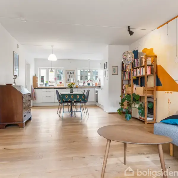 En stue med en træbord og grå sofa mod væggen. Et spisebord med stole står i det lyse åbne køkkenområde. Farverige vægmalerier og en reol med planter tilføjer personlighed.