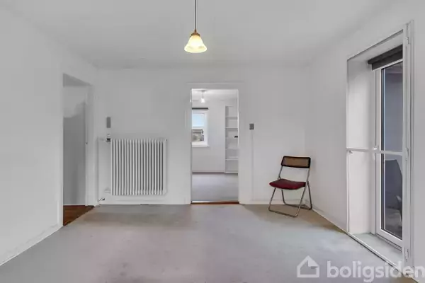 Stol står ved siden af en radiator i et hvidt rum med åbninger til andre værelser. En lampe hænger fra loftet, og en dør fører til en balkon. Ingen tekst.