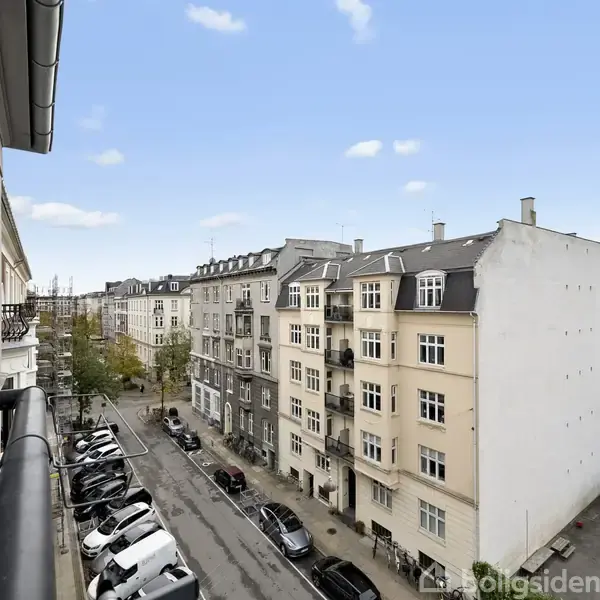 Bygningerne står langs en stille gade, hvor parkerede biler holder. Højresidede lejligheder dominerer udsigten, mens himlen er blå med få skyer.