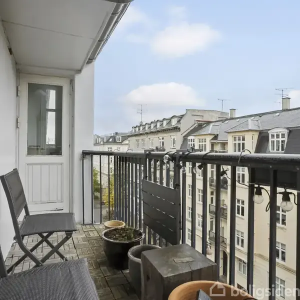 Balkonen har sorte stole og blomsterkrukker, der står stille, med udsigt over byens hustage og blå himmel. En dør leder ind til lejligheden.