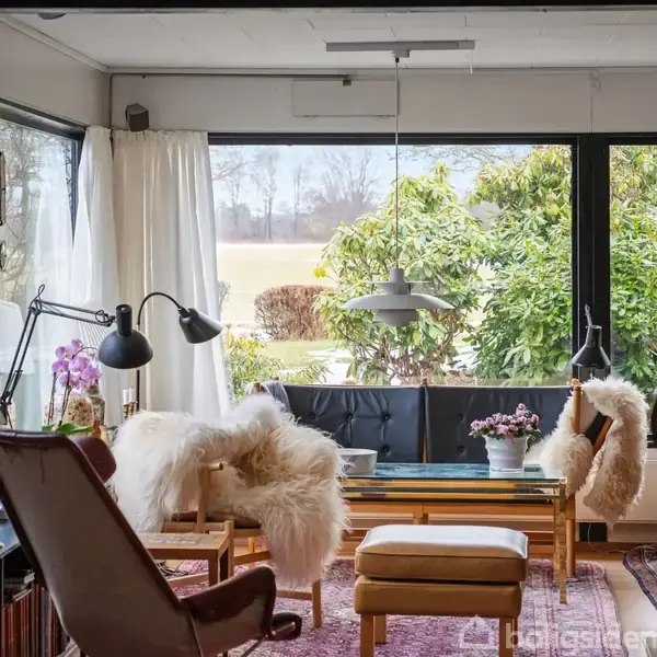 Stue med lænestole og et sofabord med blomster, omgivet af store vinduer og grønne planter udenfor. Lun og hyggelig atmosfære med behagelige tekstiler og moderne lamper.