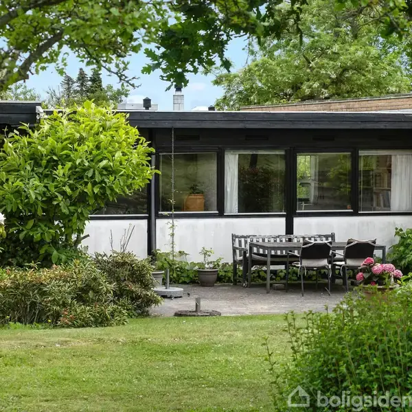 Bungalow omgivet af en stor have med buske og træer, set forfra under en lys himmel, omgivet af vinterlige træer og græsplæne i et roligt, naturskønt område.