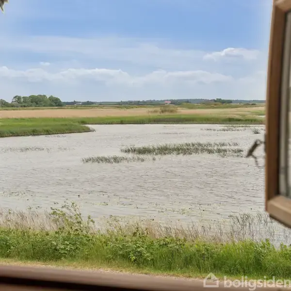 Et åbent vindue fremviser en udsigt over en rolig flod med græsarealer omkring, set fra et rum. Himlen er let overskyet.