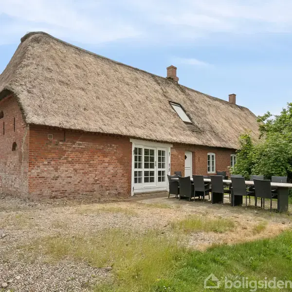 Et stråtækt murstenshus står i en grøn have med et stort udendørs spisebord. Husets facade har store vinduer og en dør, og omgivelserne er landlige.