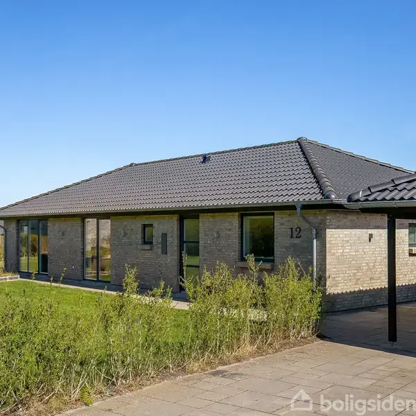 En enkeltstående, moderne murstensvilla med tagsten. Huset har store vinduer og en carport. Nummer 12 er synligt på facaden. Beliggende i et grønt område under en klar, blå himmel.
