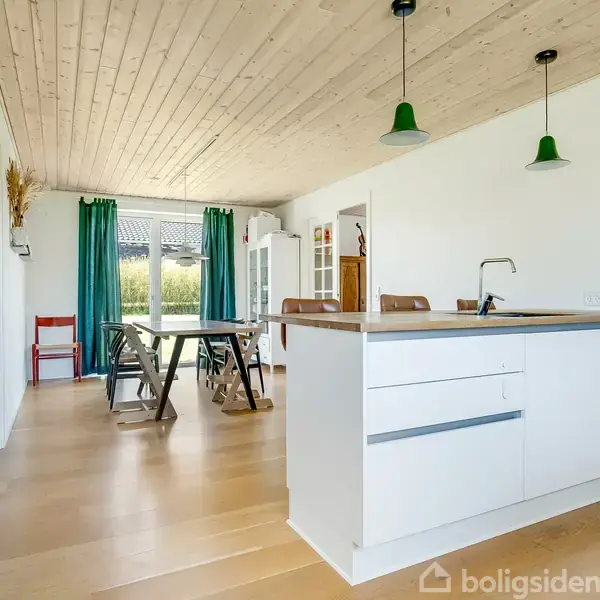 Et moderne køkken med hvidt interiør og en central ø. To grønne lamper hænger over øen. I baggrunden er der et spisebord med stole, nær et stort vindue med turkise gardiner.