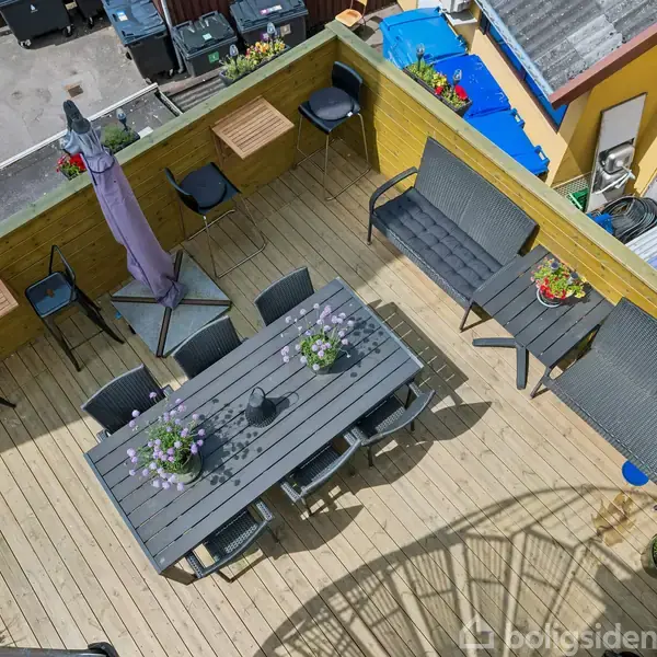 Terrasse med trædæk indeholder et langt bord med stole, blomsterdekorationer og en parasol. Bænke og små borde står langs væggen, omgivet af nabobygninger og skraldespande.