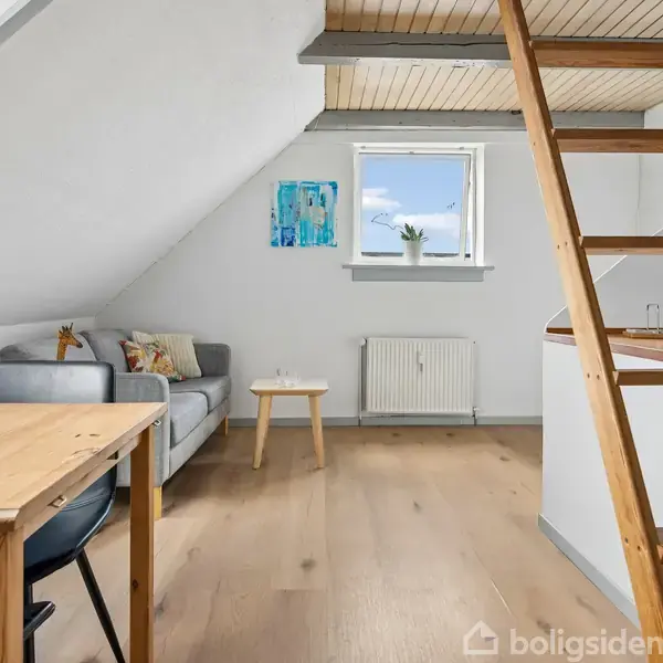 En trætrappe fører op mod et loft i en lys stue med skrå vægge. En sofa og et lille bord står ved væggen. Et vindue og en radiator er synlige.