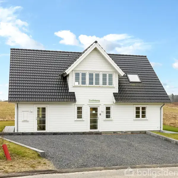 Hvidt hus med sort tag står på en grusbelagt indkørsel. Skilt over døren med teksten "Trelleborg". Omgivet af åbne marker og blå himmel med skyer.