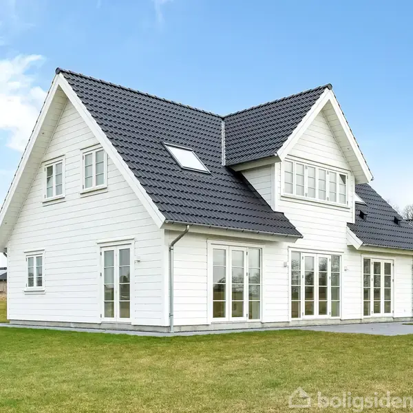 Et hvidt, to-etagers hus står på en grøn græsplæne. Det har et sort skråt tag og flere store vinduer. Baggrunden viser en åben, skyet himmel og en skov.