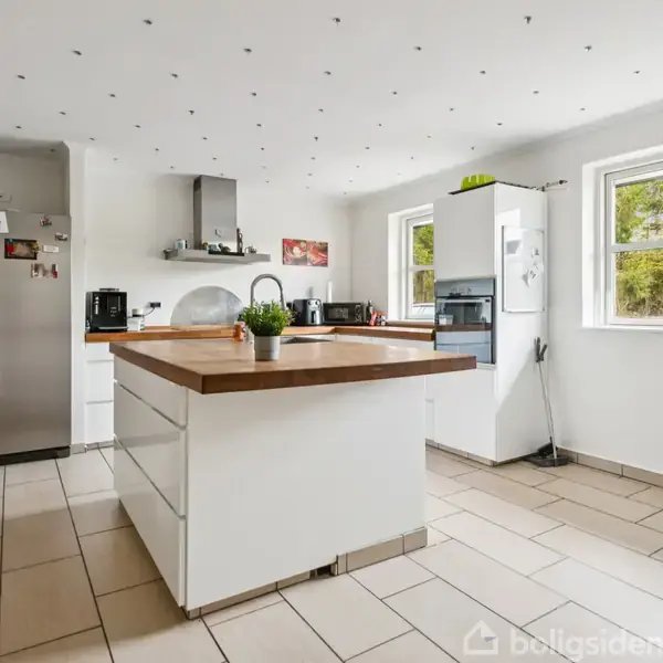 Køkken med hvid køkkenø, træbordplade og en grydeplante. Til venstre er et stort køleskab. To vinduer giver udsigt til træer udenfor. Fliser på gulvet.