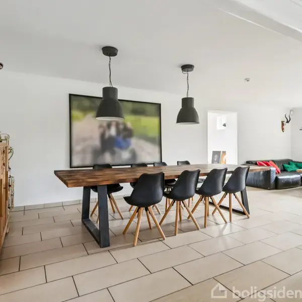 Et langt træbord står i centrum omgivet af seks sorte stole. To lamper hænger over bordet. I baggrunden ses en sofa. En hjortetrofæ hænger på væggen.