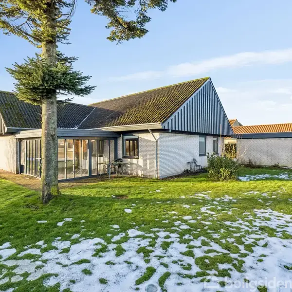 Huset står stille i en vinterhave med grøn græsplæne og let sne. Det er et hvidt murstenshus med et skråt sort tag og store vinduer. En stor træ ses i forgrunden.