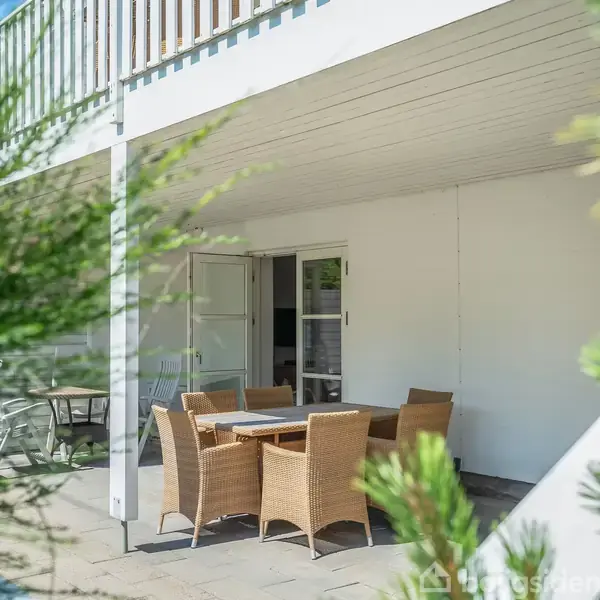 Kurvemøbler står omkring et bord på en terrasse. Døre åbner ud til rummet indenfor. Grønne planter er synlige i forgrunden, hvilket skaber en naturlig ramme.