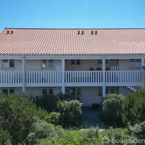 Bygning med rødt tegltag har to etager og en balkon med værn. Omkring den er der grønt buskads og en klar blå himmel over.