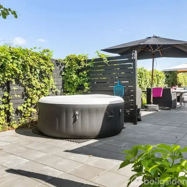 Jacuzzi placeret på en terrasse omgivet af grønne planter og sorte hegn; stole og en parasol står i baggrunden under en blå himmel.