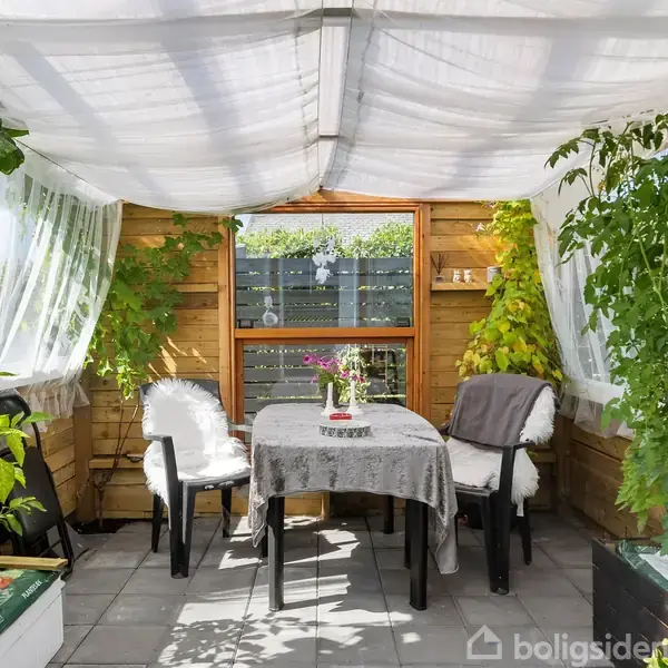 To stole står ved et bord dækket med dug og blomster. Omgivet af grønne planter, har rummet et hvidt stofloft og trægulv, hvilket skaber en hyggelig, drivhuslignende atmosfære.