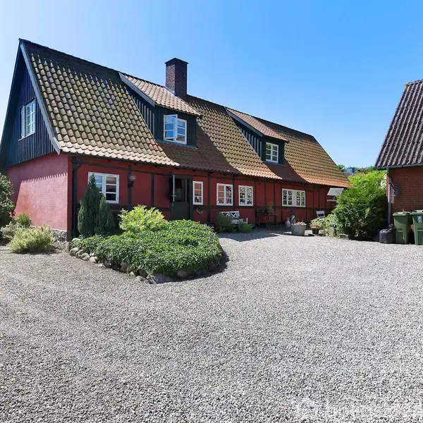 Et rødt bindingsværkshus står stille med en grusbelagt gårdsplads foran. Til venstre er der buske, og til højre ses en rød udbygning med affaldscontainere.