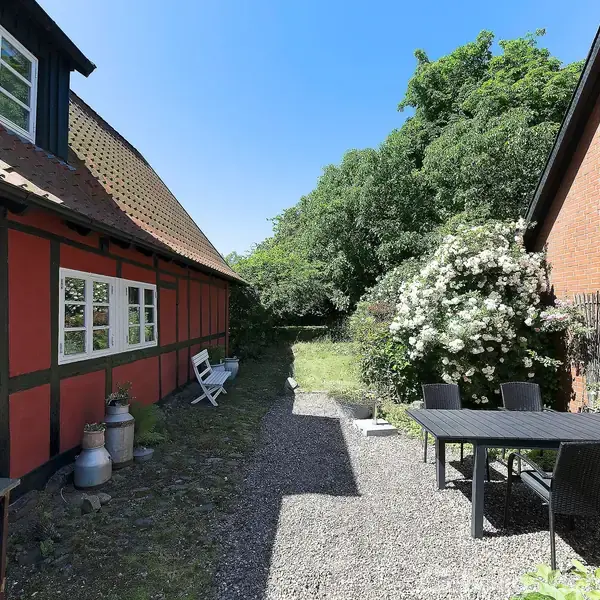 Et grusbelagt gårdhaveområde med et bord og stole. En rød bindingsværkbygning og en murstensbygning flankerer stien. Omkringliggende buske og blomster tilføjer grønt. Himlen er klar og blå.
