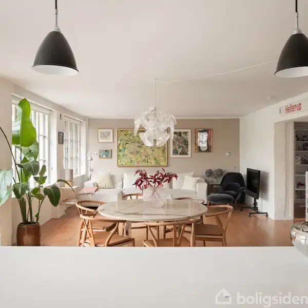Et lyst køkkenalrum med en rund spisebord med stole. En stor plante står ved vinduerne til venstre. Væggene har kunstværker og hylder. Tekst på hylde: "Hello world".
