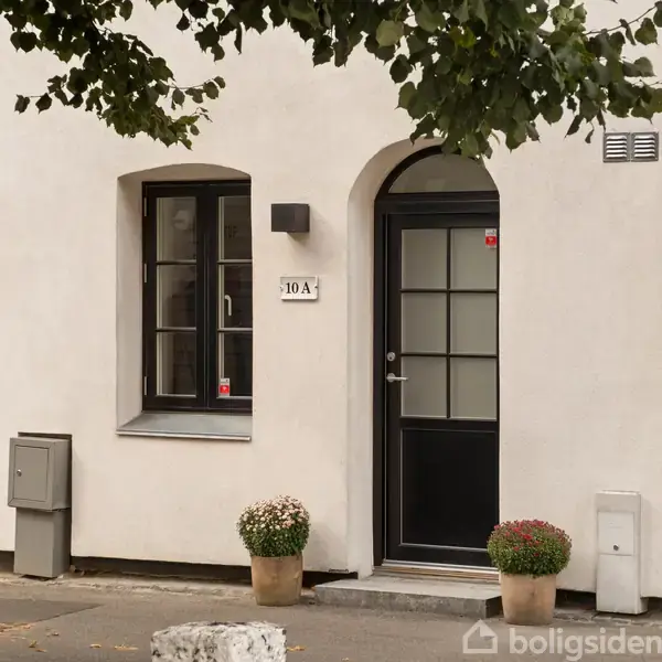 Dør med nummer "10A" er flankeret af to krukker med planter. Ved siden af døren er der et vindue og en mur med en lys på væggen. En træstamme står foran.