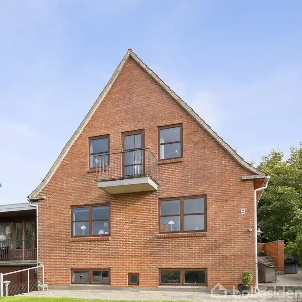 Murstenshus med spidst tag ligger i en velholdt have. Fire vinduer og en lille balkon ses på forsiden. Nummer 9 er synligt på muren. Træer omgiver huset.