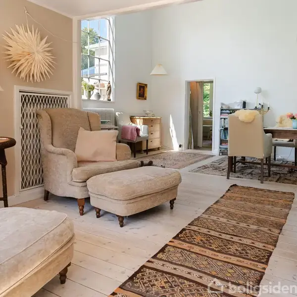Stue med beige møbler og lænestol med fodskammel, stående ved siden af et bord med lampe. Spisebord med stole i baggrunden, omgivet af lyse vægge og store vinduer.