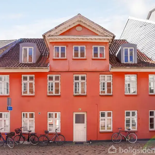 Et to-etagers, orange murstenshus med skråt tag og hvid hoveddør. Flere hvide vinduer pryder facaden. Cykler læner sig op ad huset på brostensbelagt gade.