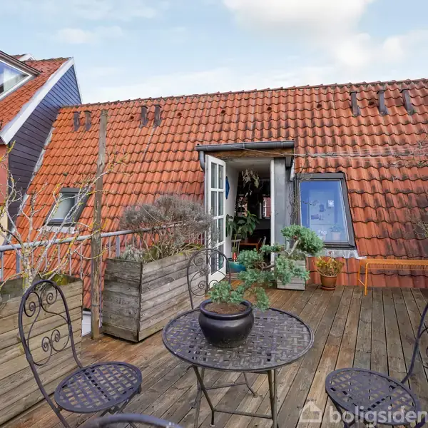 Terrasse med rundt metalbord og stole, placeret på trægulv. Baggrunden viser en bygning med rødt tegltag og trappetilgang. Planter er arrangeret i træbeholdere langs rækværket.