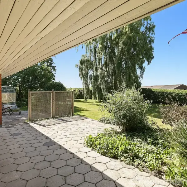 Terrasse med fliser leder ud mod en have med træer og buske. En flagstang med dansk flag står i græsset, og et langt hegn lukker området. Himlen er klar og blå.