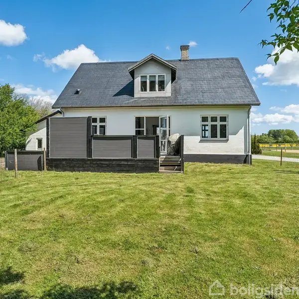 Hus med mørkt tag og hvid facade står på en velplejet græsplæne. Der er et åbent areal og træer i baggrunden under en blå himmel med skyer.