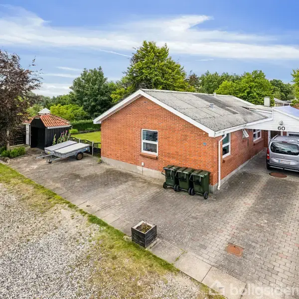 En rød murstenshus med en bil parkeret under en carport, omgivet af grønne træer og buske i forstadsområde. Husnummer "60" er synligt på carporten.