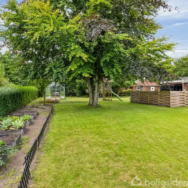 Grøntsagsbede med planter ligger i en velholdt have. Et stort træ står centralt, mens et drivhus og en legeplads ses i baggrunden ved siden af en rød bygning.
