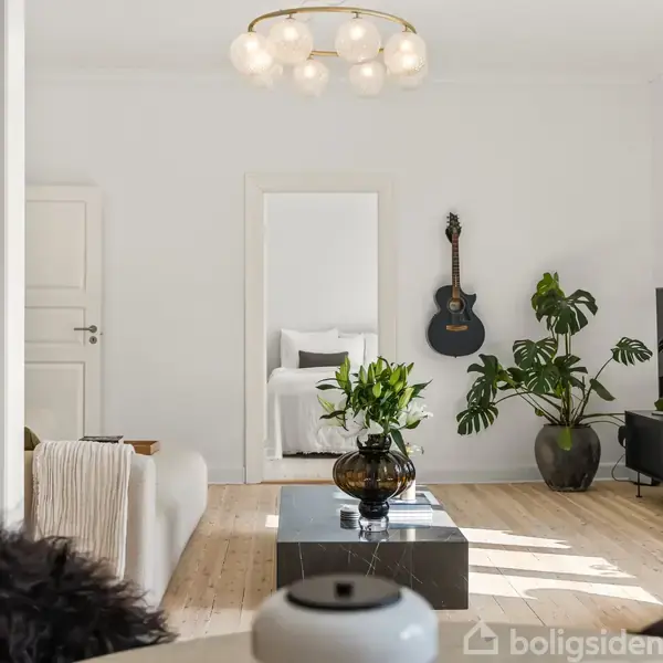 Et moderne opholdsrum med en guitar på væggen og store grønne planter. En sofagruppe står mod en åben dør, der fører til et soveværelse. En vase med blomster står på et sort bord.
