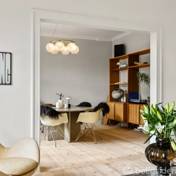 En moderne stue med lys indretning. En stor vase med planter står på sofaens bord. En reol, bord og stole ses i baggrunden. Tekst på væggen: "NOIR BLANC".