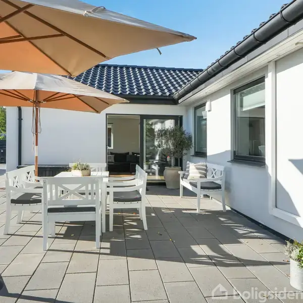 Terrasse med hvide havemøbler står under en stor parasol. Omgivet af en hvid bygning med sort tag. Grøn vegetation ses i baggrunden. En dør står åben til huset.