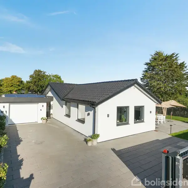 Hvidt, moderne parcelhus med sort tag står i en brolagt indkørsel. På højre side af huset er der en velplejet græsplæne med en parasol og havemøbler.