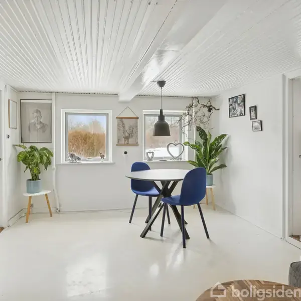 Et rundt bord med tre blå stole står i et hvidt rum med billeder på væggene. En sort lampe hænger fra loftet. To planter står ved vinduerne og det er dagslys.