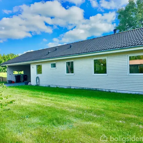 En hvid villa står stille med grå tag i en grøn have, omkranset af buske og træer under en blå himmel med skyer.