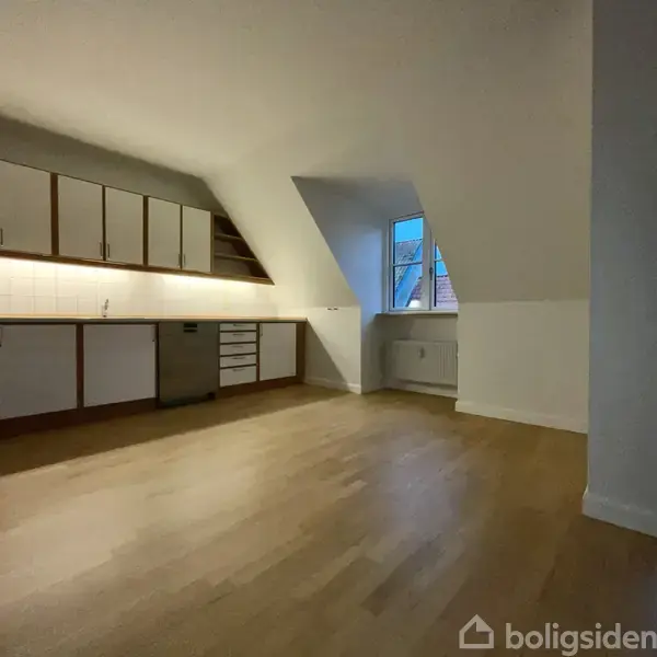 Et køkken med hvide skabe og indbygget ovn står stille. Rummet er tomt med trægulv og et skråvindue, der slår lys ind, omgivet af et rent, minimalistisk interiør.