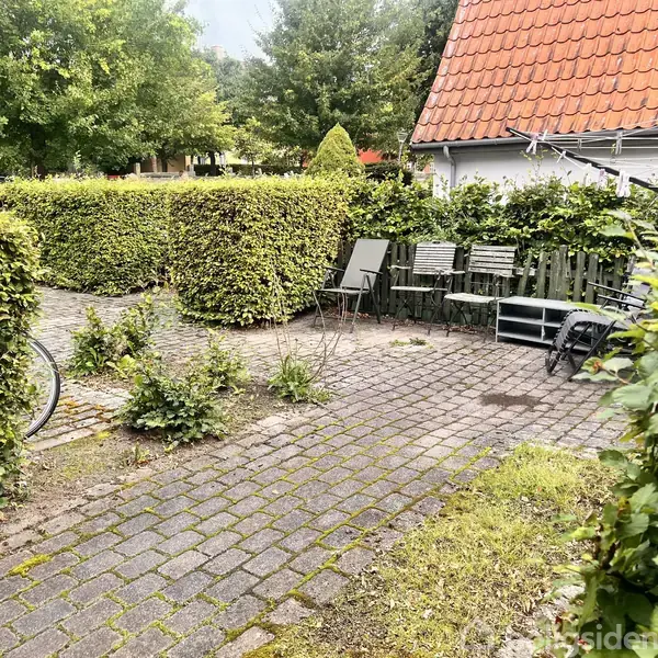 En flisebelagt gårdhave med havemøbler og cykel, omkredset af trimmede hække. I baggrunden ses hus med rødt tegltag og en række træer.