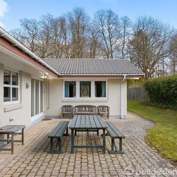 Et træbord med bænke står på en flisebelagt terrasse. I baggrunden er der et hus med store vinduer og en græsplæne flankeret af buske og træer.