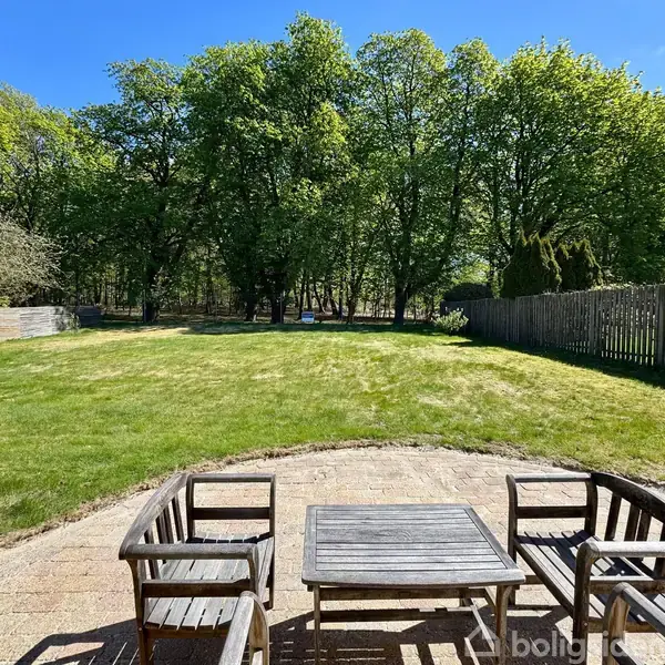 To træstole og et bord står på en flisebelagt terrasse. Baggrunden består af en stor græsplæne omgivet af træer og et hegn. Boligsiden-vandmærke ignoreres.