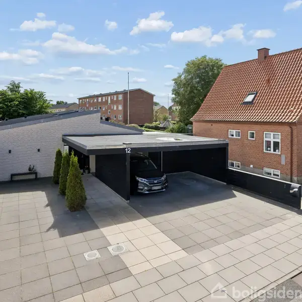 Carport med sort bil parkeret, omgivet af betonfliser og grønne buske. Ved siden af ses en rød murstensbygning med rødt tag. Nummeret "12" er synligt over carporten.