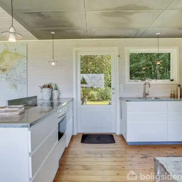 Lyst køkken med hvide skabe og træbordplade, stående i et rum med trægulv. Et verdenskort hænger på væggen, og der er udsigt til haven gennem en glasdør.