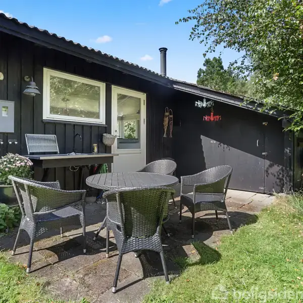 Et rundt bord med fire kurvestole står på en terrasse foran et sortmalet træhus. Terrassen er omgivet af grønt græs og planter. En skygge falder på husets væg.