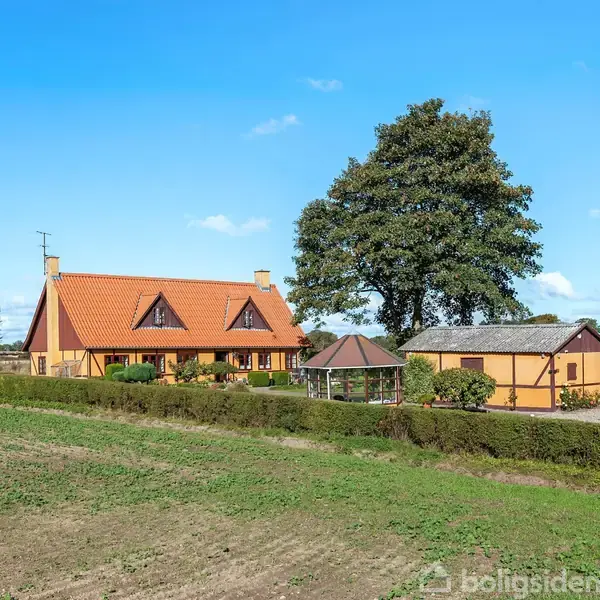 Et gult bindingsværkshus med rødt tegltag står omgivet af grønne buske på en landlig ejendom. En stor træ i baggrunden mod en blå himmel. Ingen tekst til stede.