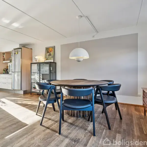 Et rundt spisebord med sorte stole står i en lys spisestue. En stor lampe hænger over bordet. I baggrunden ses et køkken og et vitrineskab.