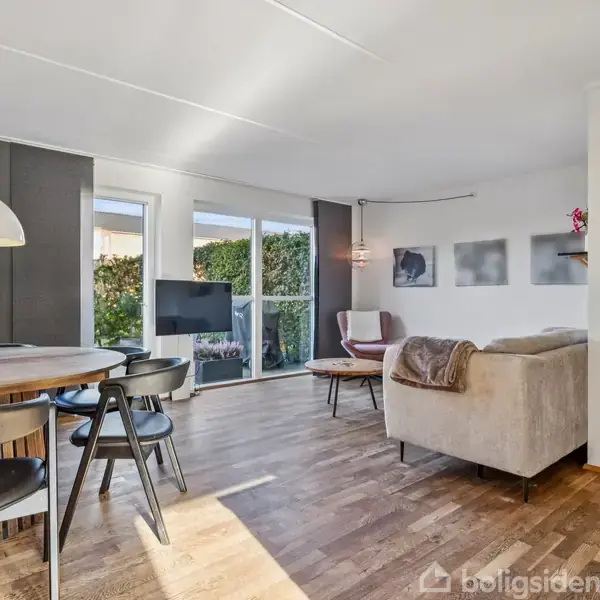 En stue med en lysegrå sofa ved trappen, et rundt spisebord med sorte stole til venstre, og et tv ved store vinduer, som giver udsigt til en have.