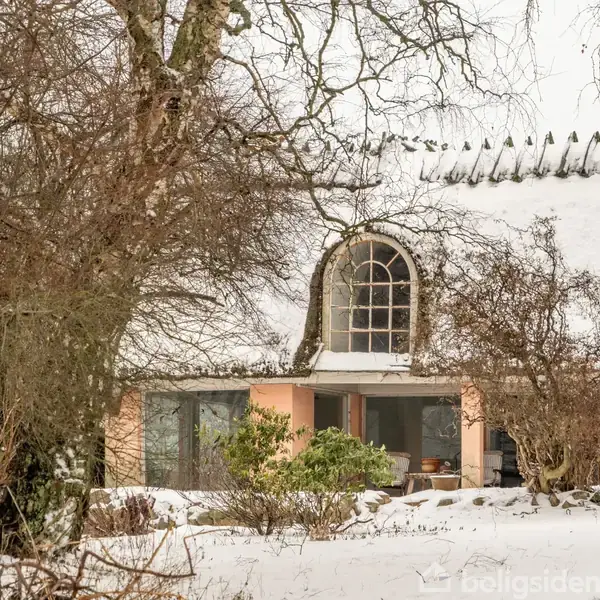 Bindingsværkshus dækket af sne, omgivet af nøgne træer. Sneen skjuler tagets struktur delvist, mens en stor bueformet vinduesramme centreres i midten. Vinterpræget landskab skaber en rolig atmosfære.